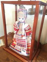 Front and side view of the porcelain doll in a wooden-framed glass case showing detailed traditional attire and display setup.