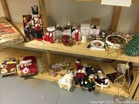 Wide shot of various Christmas decorations and figurines on wooden shelves showing assortment including boxed ornaments, Nutcracker soldier, Santa-themed gift boxes, and ceramic Christmas tree lamp.