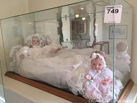 Full view of glass display case containing two vinyl dolls, 'Rose' doll seated in pink and white, other doll reposed in white baptism dress.