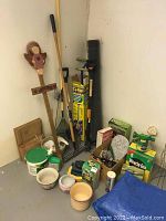 Wide shot showing collection of garden tools including rakes, shovel, broom, plant pots, gardening supplies and decorative wooden stake with angel figure.