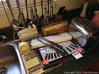 Kitchen counter with Ginsu knife set box, wooden cheese boards, yellow egg timer, metal utensils on hanging rack, handheld mixer, and stainless steel toaster.