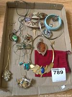Overview of assorted jewelry in cardboard box including necklaces, bracelets, and brooches, with large amber brooch visible on red pouch.