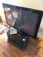 Front view of Samsung Plasma TV on JVC stand, showing TV screen, remote, and user guide on stand.