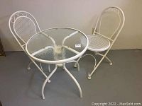 White metal bistro set showing round glass top table and two matching folding chairs with mesh seats and backs.