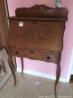 Full view of wooden secretary desk standing against a pink wall, showing elegant curved legs and top carved panel.