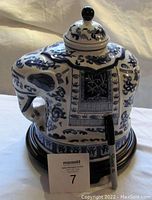 Front and side view of the blue and white ceramic elephant-shaped ginger jar sitting on a polished wooden base, with a MaxSold lot number card and marker for scale.