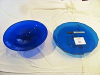 Both cobalt blue footed cake plates side by side on a white cloth background, showing size and shape difference alongside a marker for scale.