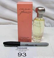 Front view of Estee Lauder Pleasures Eau De Parfum bottle next to its pink box and a Sharpie pen for scale. Bottle is clear glass with silver cap, liquid inside is pale yellow. Box shows product name and branding.