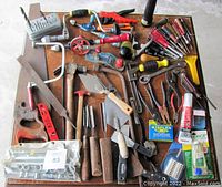 Wide view of assorted hand tools laid out on a flat surface, showing vintage hand drills, chisels with wooden handles, hand saws, pliers, screwdrivers, and various other small hand tools with accessories such as glue tubes and staple packs.