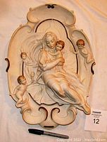Full view of the porcelain wall plaque showing Madonna with baby Jesus and cherubs, along with pen and card for scale.