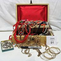Full view of the open gold-tone jewelry box with red satin interior and various colorful costume jewelry spilling out.
