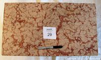 Top view of rectangular pink and light brown mottled marble slab surface of the table with a marker for scale and a lot number card.