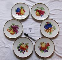 Six dessert plates arranged in two rows on white surface, showing different fruit designs and gold rims, with marking card and pen nearby.