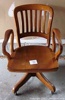 Front view of the vintage Krug style banker's chair showing vertical slat backrest, wooden armrests, and solid wooden seat with minor split.