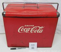 Front view of red Coca-Cola metal cooler with white script logo 'Drink Coca-Cola' and metal handle folded down.