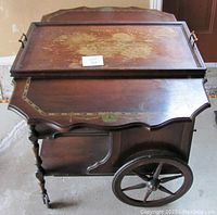 Front and top view of the vintage wooden tea cart showing the drop leaf extension closed, decorative golden accents and floral design on the top wooden surface, and the large wooden wheel with small caster on the opposite side.