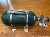 Green cylindrical aircraft oxygen tank with metal mounting brackets, pressure gauge and valve, and Sky Ox label.