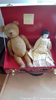 Photo showing stuffed toy bear and doll in yellow dress placed in a red attic storage box with label.
