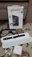 Photo showing black rubber Apple Watch band in plastic case, single white Apple AirPod in small box, coiled USB charging cables, and boxed screen protector for iPad Mini 3