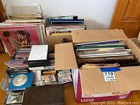 Three boxes with vinyl records in sleeves and assorted CDs, cassettes, DVDs and VHS tapes on floor.