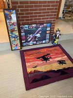 Lot items arranged on floor against brick wall: framed vertical textile art with jets, framed Christen Eagle II airplane poster, textile wall hanging with silhouette biplanes, aviator statue on floor