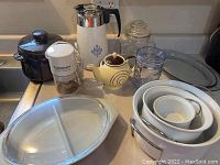 Photo showing assortment of kitchen items including black lidded brown ceramic pot, Cuisinart white chopper, white Corningware coffee pot with blue floral design, hand painted yellow ceramic teapot, clear glass storage jar, plastic storage jar, Corningware roaster with lid, and nested white porcelain bowls with handles.