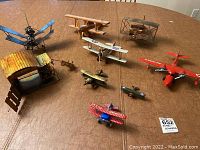 Nine decorative airplane models shown on a table including wooden hangar structure, various biplanes, monoplanes in different colors and materials