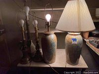 Two tall ceramic base lamps on a table, one with a cream-colored pleated lampshade, and three brass base lamps behind them, showing the full set of five lamps.