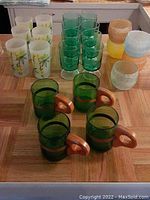 Full view of 6 frosted bamboo motif highball glasses, 8 greenish-blue short stemmed glasses, 4 green bottle mugs with wooden handles, and 5 rounded rubber-coated rocks glasses on a wood patterned surface.