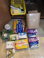 Under sink storage bin with sponges, cleaning wipes, garbage bags, wraps, plastic utensils, and a wastebasket on floor