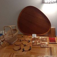 Overall lot view showing an egg-shaped wooden tray, round wooden tray with rim, wooden coasters with animal figures and inns illustrations, boxed set of red-handled olive forks, and woven wine bottle basket.