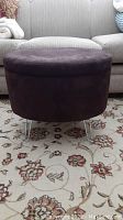 Closed view of plum-colored oval design ottoman on silver hairpin legs in front of a beige sofa on a floral carpet.