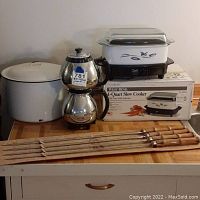 Full set of listed kitchen items including enamel stockpot, slow cooker on box, coffee pot and skewers laid out on table
