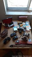 Overview of the vintage cameras, binoculars and accessories arranged on a wooden surface near a window. Visible cameras include Minolta, Fuji, Edinex and Kodak models along with binoculars, camera straps, flash bulbs, and a leather case.