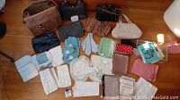 Photo showing assorted vintage purses, handbags, wallets, and array of linens including napkins, scarves, and doilies spread on hardwood floor.