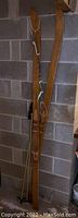 Pair of wooden skis leaning against a concrete wall, showing full skis and part of a ski pole