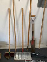 Photo showing six yard tools against a white wall including a push broom, a small hand broom, metal snow shovel, metal rake, post hole shovel, and garden hoe all with wooden handles.