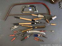 Various yard and garden hand tools laid on dark surface: three saws, several pruners, trimmers, trowels, and a bulb planting tool visible