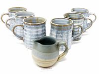 Six blue drip glaze pottery mugs arranged behind a matching pottery creamer with beige and blue shades on a white background.
