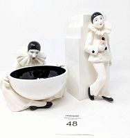 Two Pierrot clown ceramic figures: one holding a black and white bowl seated, the other standing by a tall white geometric ceramic vase holding a pink rose.