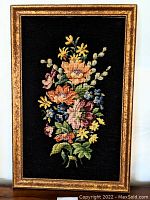Full view of the framed needlepoint depicting vibrant flowers on a black backdrop with a gold textured frame.