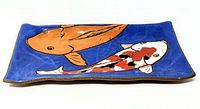 Side angle of rectangular hand painted pottery dish featuring orange and white koi fish on blue background, showing slight edge curvature.
