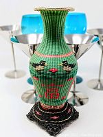 Front angle view of vintage Chinese woven vase with teal wine glasses and stainless steel martini goblets behind it.