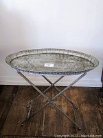 Front view of the oval silver folding side table standing open on crossed folding legs.