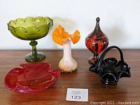 Grouping of six vintage Mid Century art glass and ceramic decorative pieces on neutral background, including an olive green pedestal dish, orange ruffled vase, red dish, black basket with handle, red and black jar, and white/orange vase