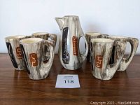 All items on table showing the coffee pot with its distinctive abstract hand-painted design, surrounded by six matching mugs with sculptural handles and coordinating patterns.