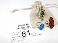 Three rings placed near drawstring linen bag, showing blue topaz, orange jasper and mystic topaz with green accent stones, sizes noted on paper