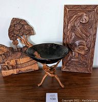 Photo shows the full lot with carved wooden tripod stand holding a dark carved bowl and two carved wall hangings in tribal style.
