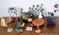 Overview of vintage metal, ceramic, and wooden collectible decor items showing peacock, turtles, penguin, carved wood items and metal sculptures on display surface.