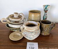 Photo showing group of nine vintage pottery pieces with different rustic glazes including vessels, mugs, candle holders, and bowls arranged on a wooden surface.